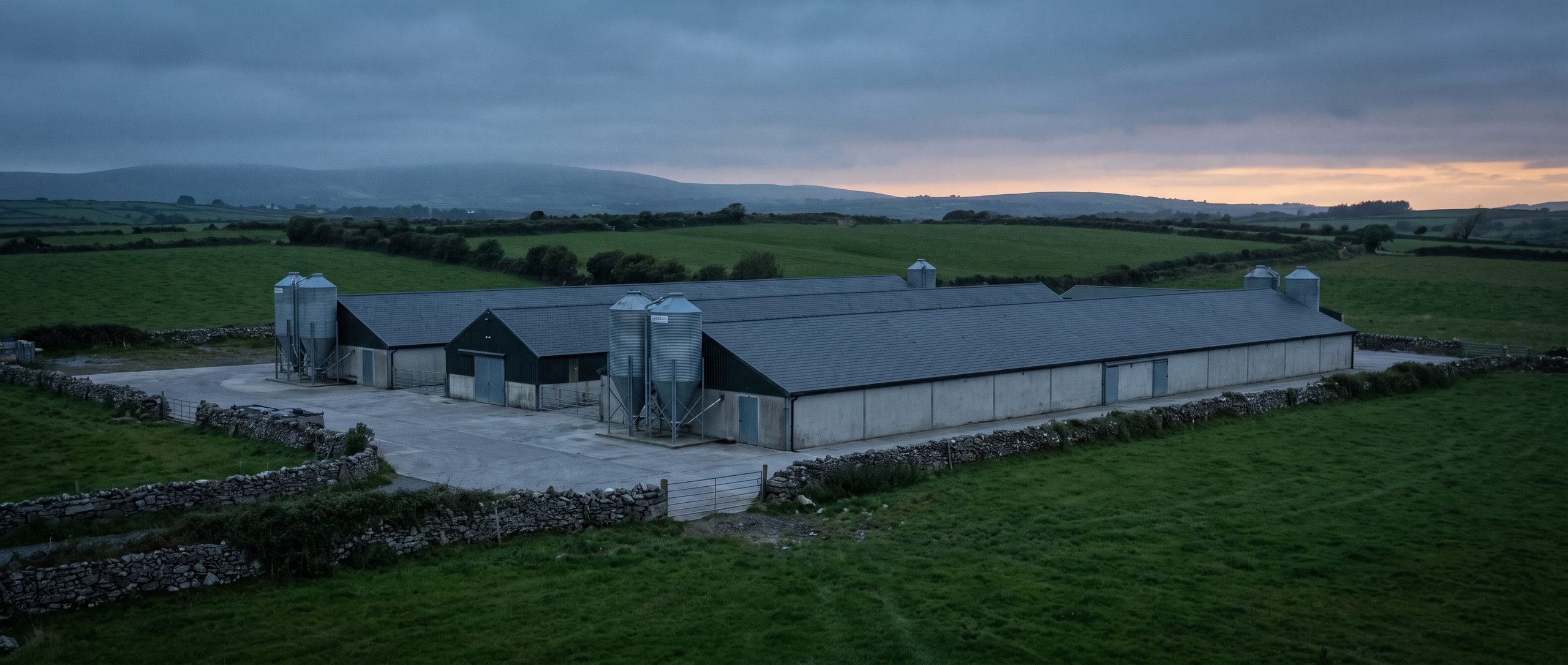 The Kilfilum pig unit at dusk: slate-roofed piggeries, four silver feed silos and a concrete yard, walled by dry-stone banks in the County Clare countryside.